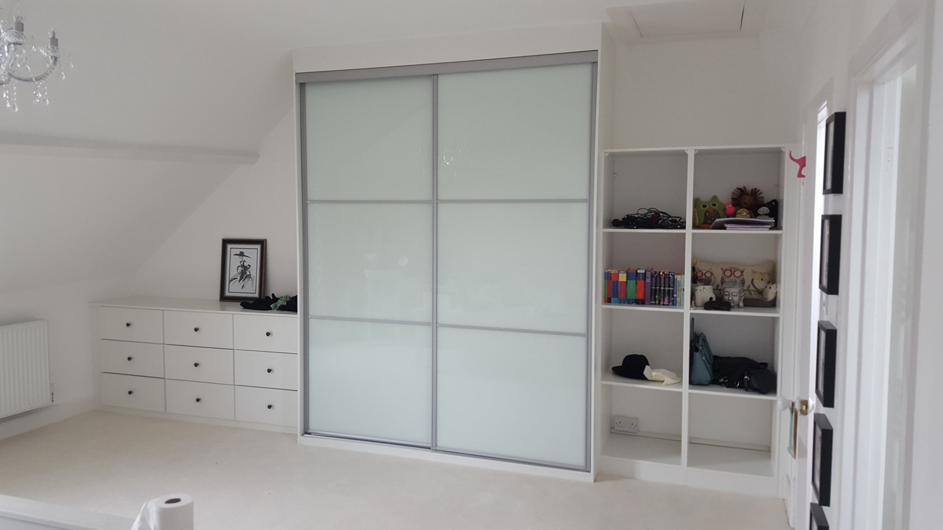 Two pale coloured glass sliding doors with silver frames and horizontal divisions, beside open white shelving and matching drawers under a sloping ceiling in Carmarthen