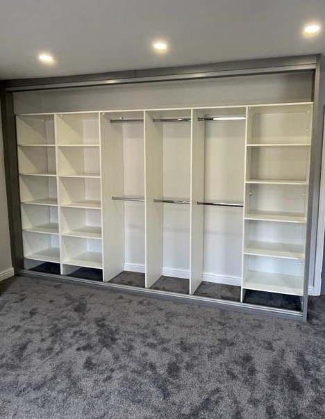 Inside of a fitted sliding wardrobe run with the doors removed, showing white shelf columns and hanging rails at two heights